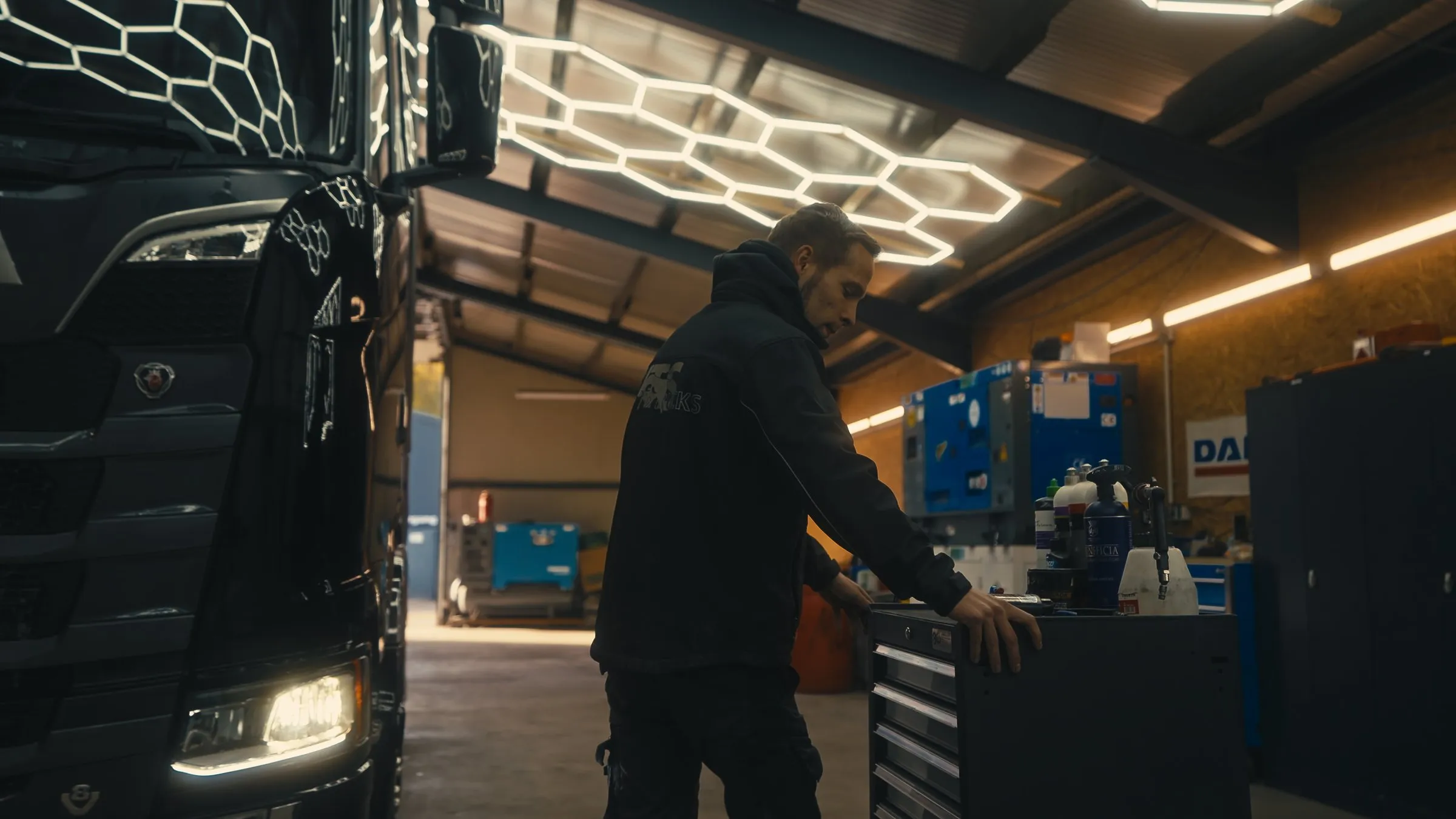 Mechaniker am Werkzeugwagen in der Werkstatthalle, daneben ein Scania mit eingeschaltetem Licht